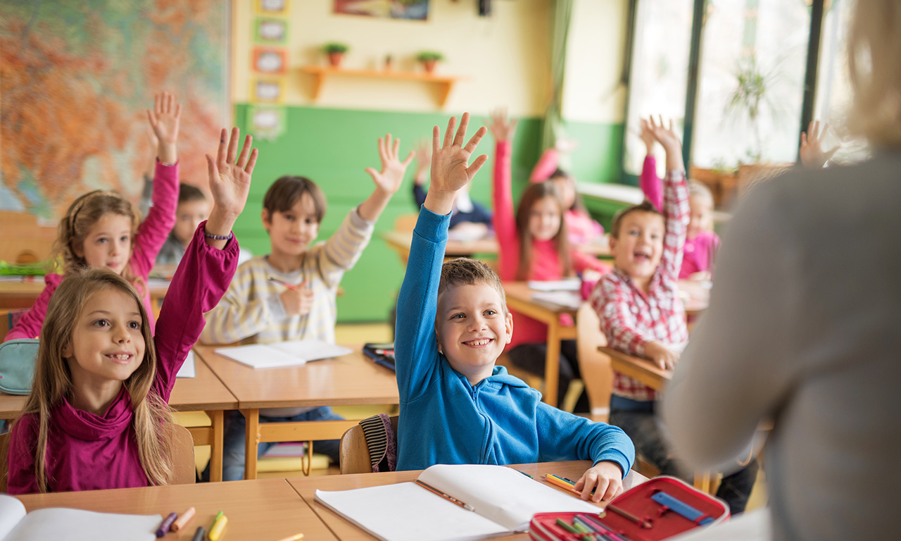bambini della prima classe primaria alzano la mano per rispondere alla maestra
